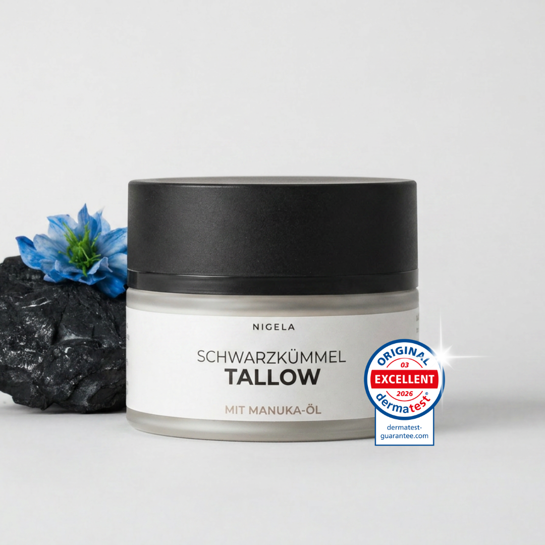 Nigela Schwarzkümmel Tallow neben schwarzem Stein und blauer Schwarzkümmelblüte auf hellem Hintergrund.